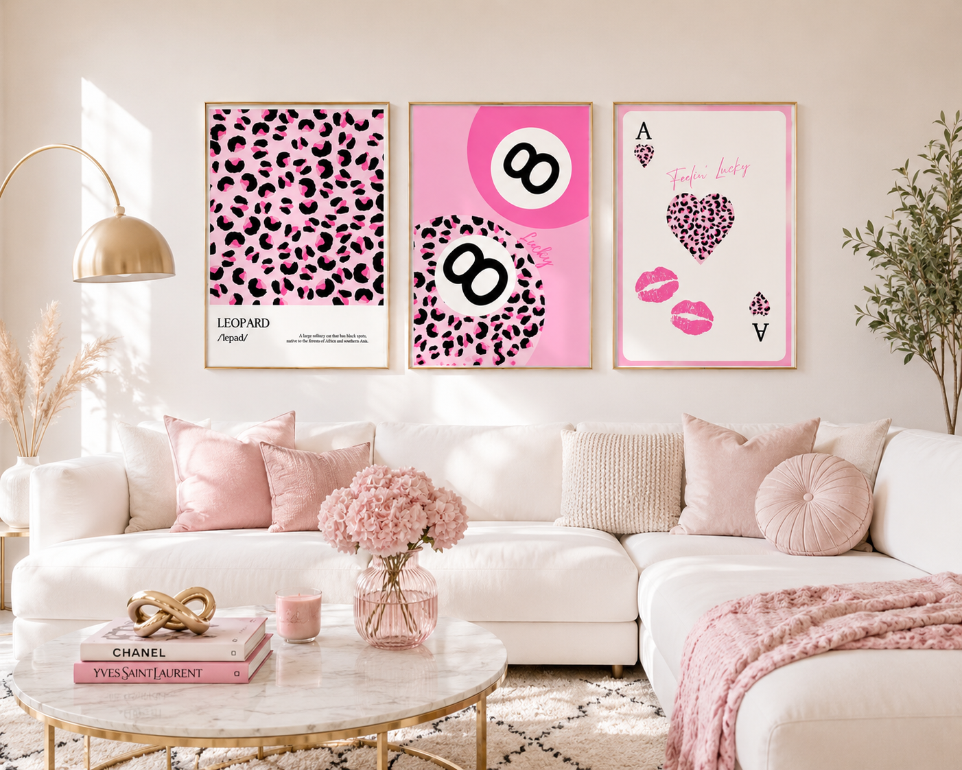 Three framed pink leopard-print and graphic wall art prints displayed above a white sectional sofa in a styled living room. The prints feature leopard patterns, an eight-ball motif, and playing card graphics with pink lip prints.