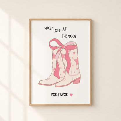 A framed wall art print featuring a pink cowboy boot illustration with the text 'SHOES OFF AT THE DOOR' and 'POR FAVOR' below, hanging on a light beige wall with soft window-shadow lighting.