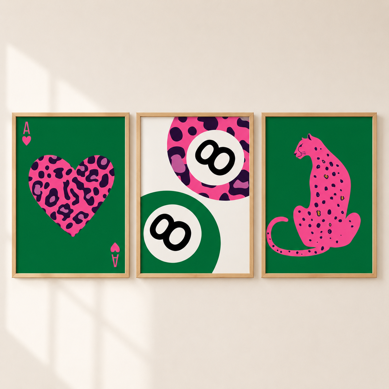 Three framed wall art prints with a green background and bold pink graphic designs hang on a beige wall. The left print shows an ace of hearts with a pink leopard-print heart; the center print features two white 8-balls over pink-and-purple leopard and green circular patterns; the right print shows a pink leopard sitting in profile with black spots.