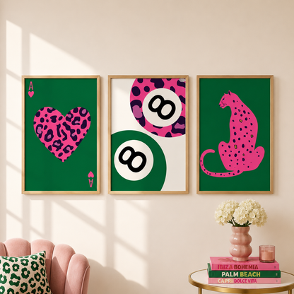 Three framed wall art prints with bold pink leopard-style graphics are displayed on a beige wall in a styled room. The left print shows a heart-shaped ace of spades, the center shows stacked 8-balls, and the right shows a leopard cat silhouette.