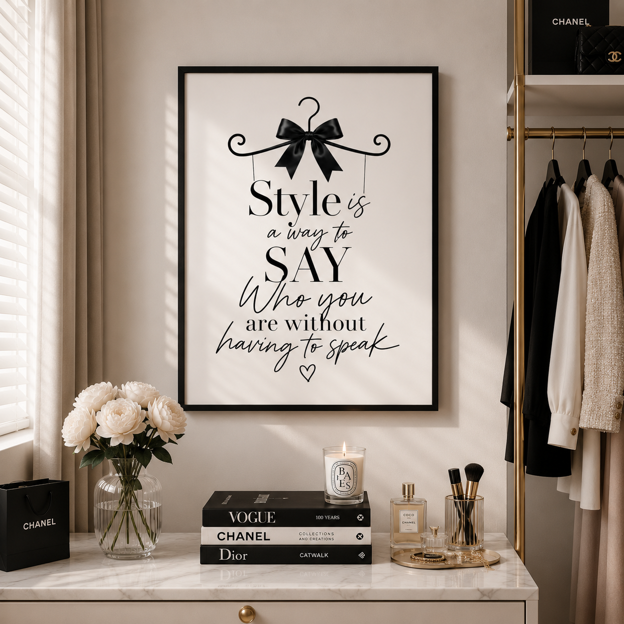 A framed wall art print with a black border and black typography reading 'Style is a way to SAY Who you are without having to speak' hangs on a beige wall in a styled interior. The room includes a marble console table with flowers, books, candles, and perfume, and a clothing rack with garments.