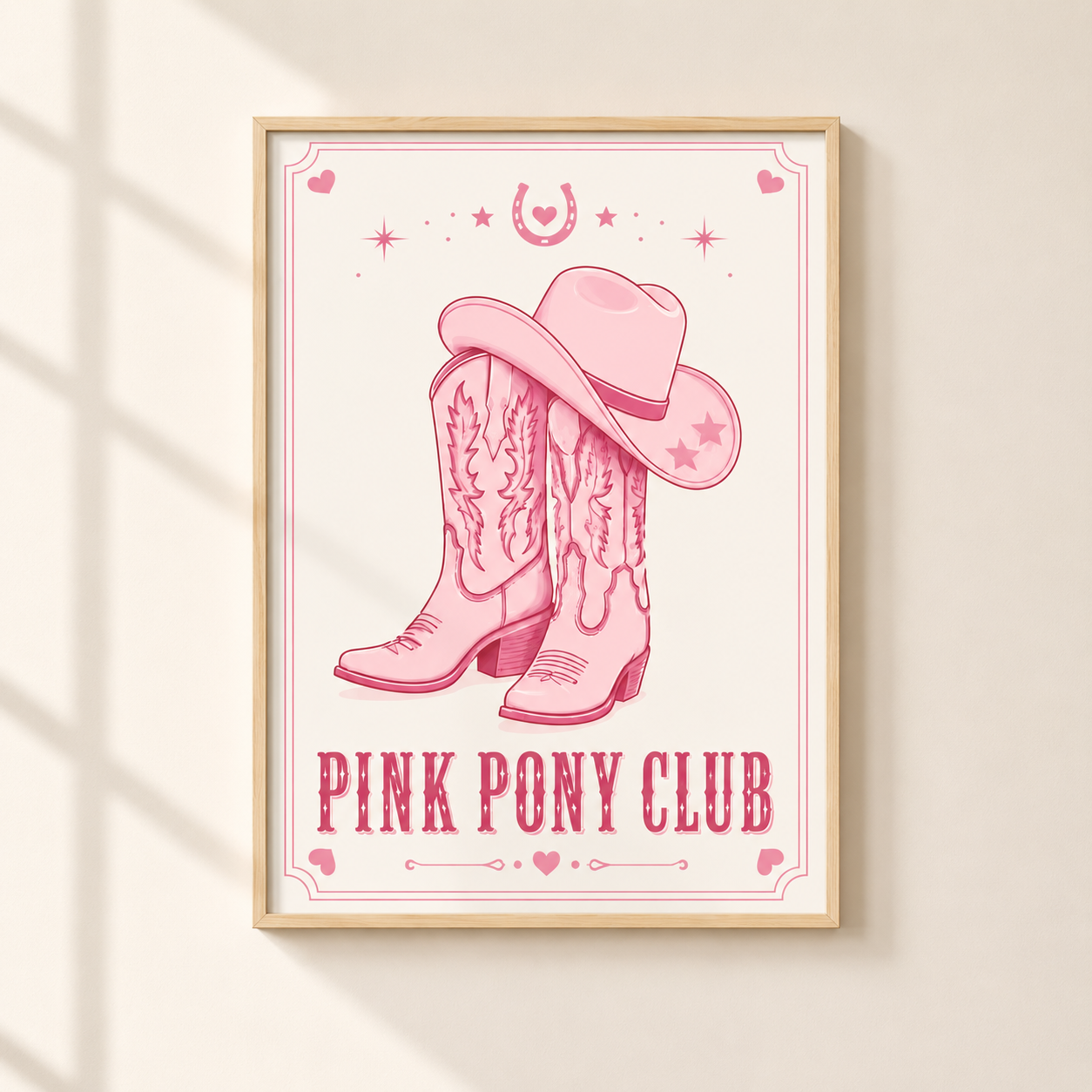 A framed art print featuring a pink cowboy hat and boots design with the text 'PINK PONY CLUB' on a cream background, hanging on a light beige wall with soft window shadow patterns.
