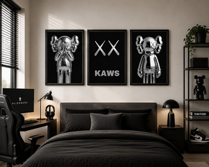 A three-piece framed wall art set featuring metallic silver cartoon bear figures and the text 'XX KAWS' on a black background, displayed above a bed in a modern black-themed bedroom.