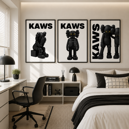 A styled bedroom featuring three framed black-and-white 'KAWS' art prints above a bed. The prints show black character figures with yellow X eyes, and the room includes a desk, chair, lamps, and neutral bedding.