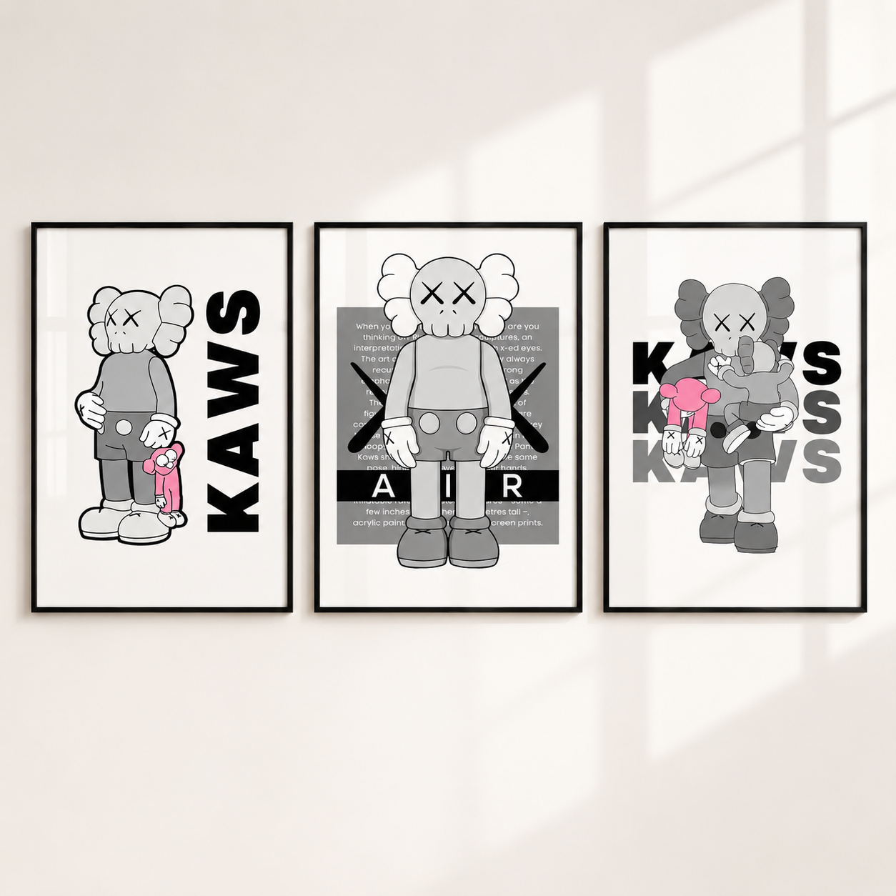 Three framed wall art prints featuring stylized KAWS character illustrations in black, white, and gray, with one print including a small pink plush figure, displayed on a light wall.