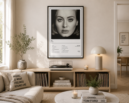 A framed black and white portrait poster of Adele is displayed on a beige wall in a modern, sunlit living room above a wooden console table.