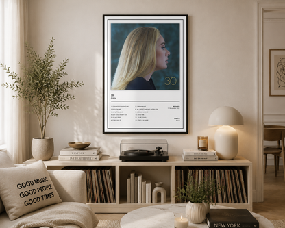 A framed vertical music poster of Adele's 30th anniversary tour hanging on a beige wall in a modern, styled living room with a bookshelf, lamp, and plants.
