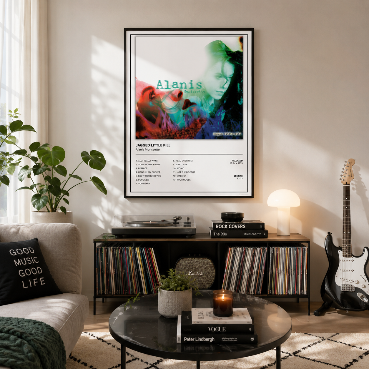 A framed music poster of Alanis Morissette's 'Jagged Little Pill' hanging on a beige wall in a modern, sunlit living room. The room features a black record store cabinet filled with vinyl records, a black coffee table with books, a green sofa with a decorative pillow, and a black electric guitar leaning against the wall.