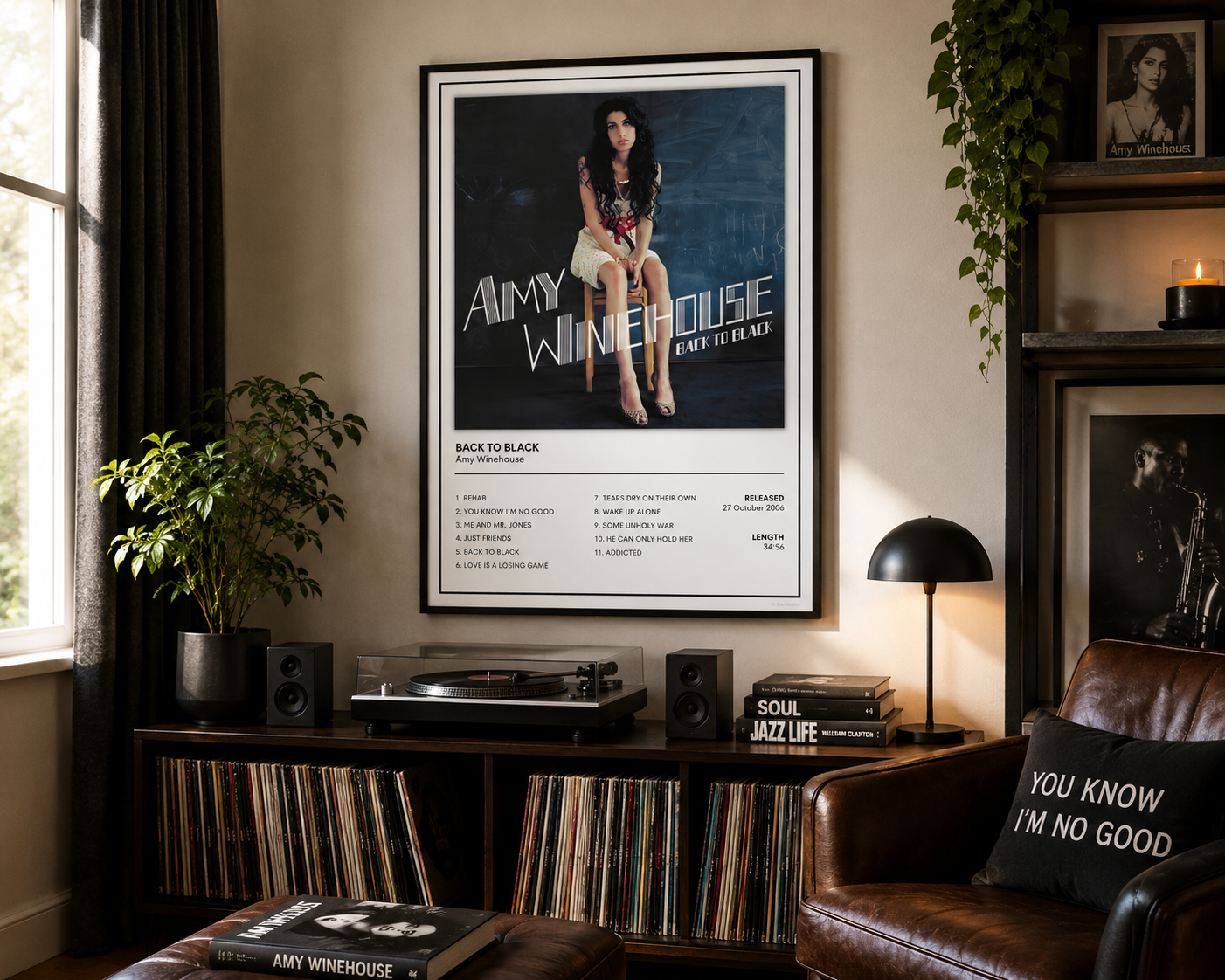 A framed Amy Winehouse 'Back To Black' poster hanging on a beige wall in a styled living room above a record shelf, with a leather armchair, lamp, and plants nearby.