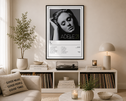 A framed black and white Adele 21 album poster hangs on a beige wall in a styled living room setting above a white record shelf.
