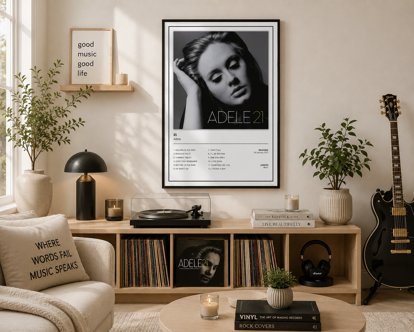 A framed black-and-white Adele 21 album poster hangs on a beige wall above a wooden media console in a styled living room. The room includes a chair with a quote pillow, potted plants, a turntable, headphones, a guitar, and candles.