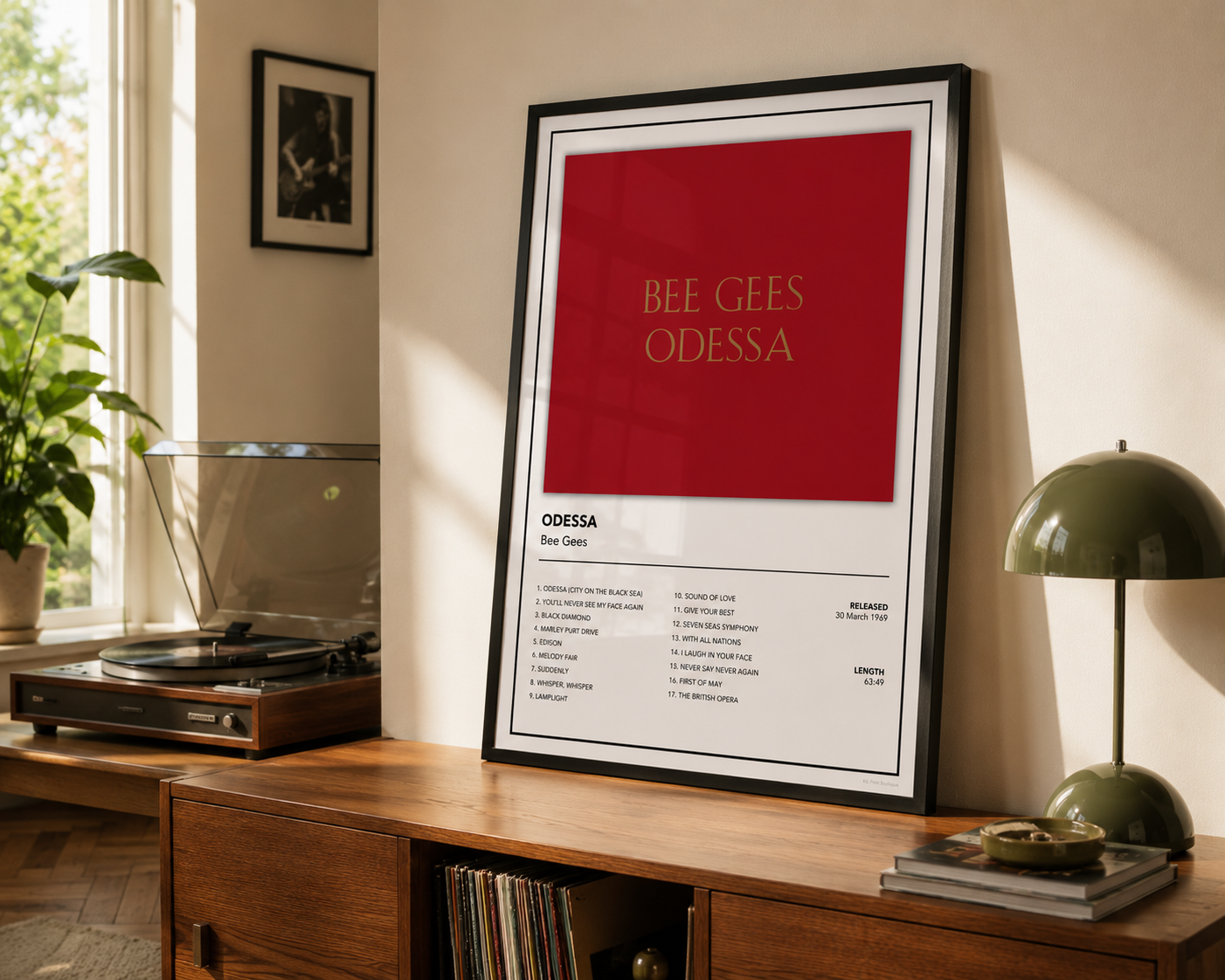 A framed music poster for the Bee Gees' 'Odessa' leaning on a wooden console table in a sunlit room, with a record player, plant, and green table lamp nearby.