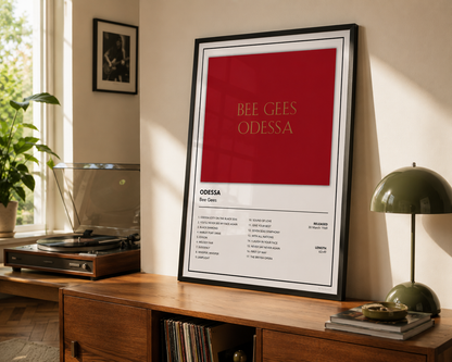 A framed music poster for the Bee Gees' 'Odessa' leaning on a wooden console table in a sunlit room, with a record player, plant, and green table lamp nearby.