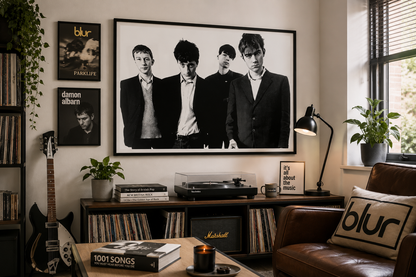 A large black and white framed poster of the band Blur hangs on a beige wall in a styled living room. The room features a brown leather sofa with a matching pillow, a wooden record player, a guitar, and other framed Blur album art.