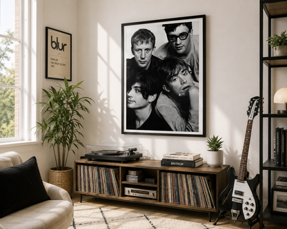 A large black and white framed poster of the band Blur hangs on a light beige wall in a modern, sunlit living room. The room features a wooden media console filled with vinyl records, a record player, a potted plant, a guitar, and a sofa in the foreground.