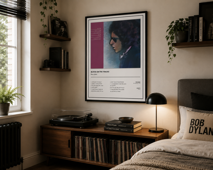 A framed Bob Dylan 'Blood on the Tracks' poster hanging on a beige wall in a styled bedroom setting with a wooden console, lamp, and bed.