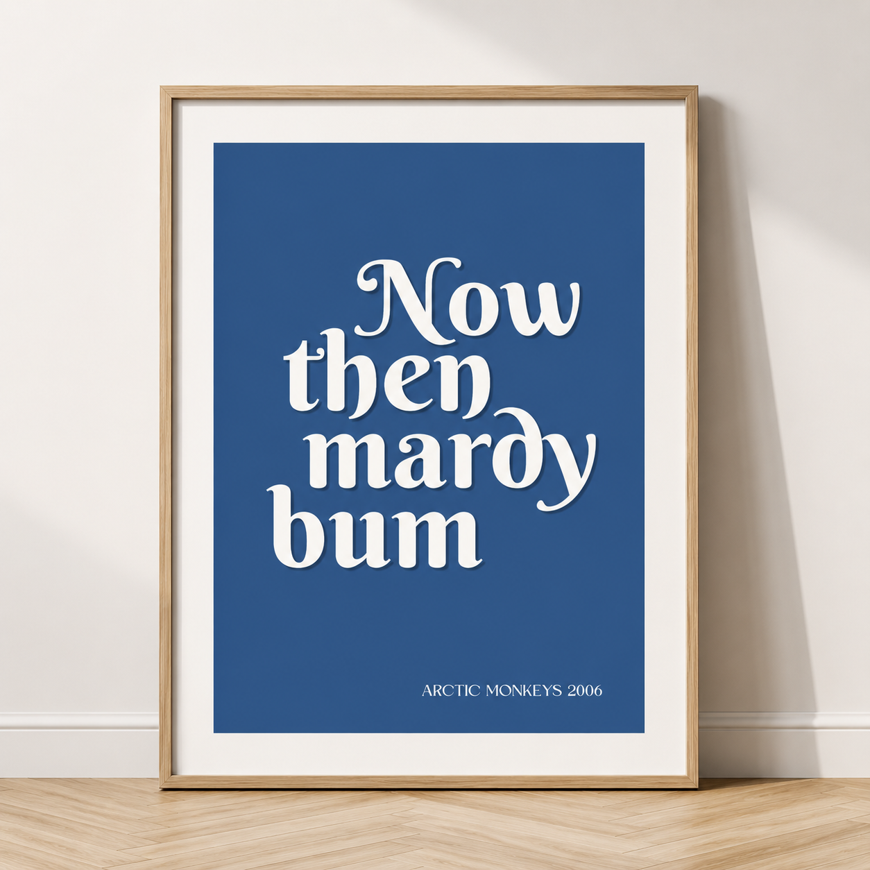 Blue Arctic Monkeys "Now Then Mardy Bum" typography wall art displayed above a bed in a natural oak frame and white mount.