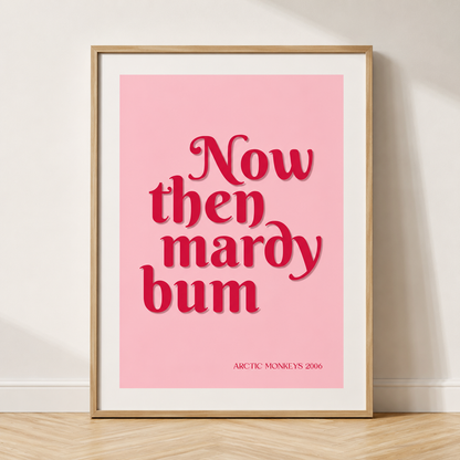 Pink Arctic Monkeys "Now Then Mardy Bum" typography print in a natural oak frame with a white mount against a minimalist neutral wall.