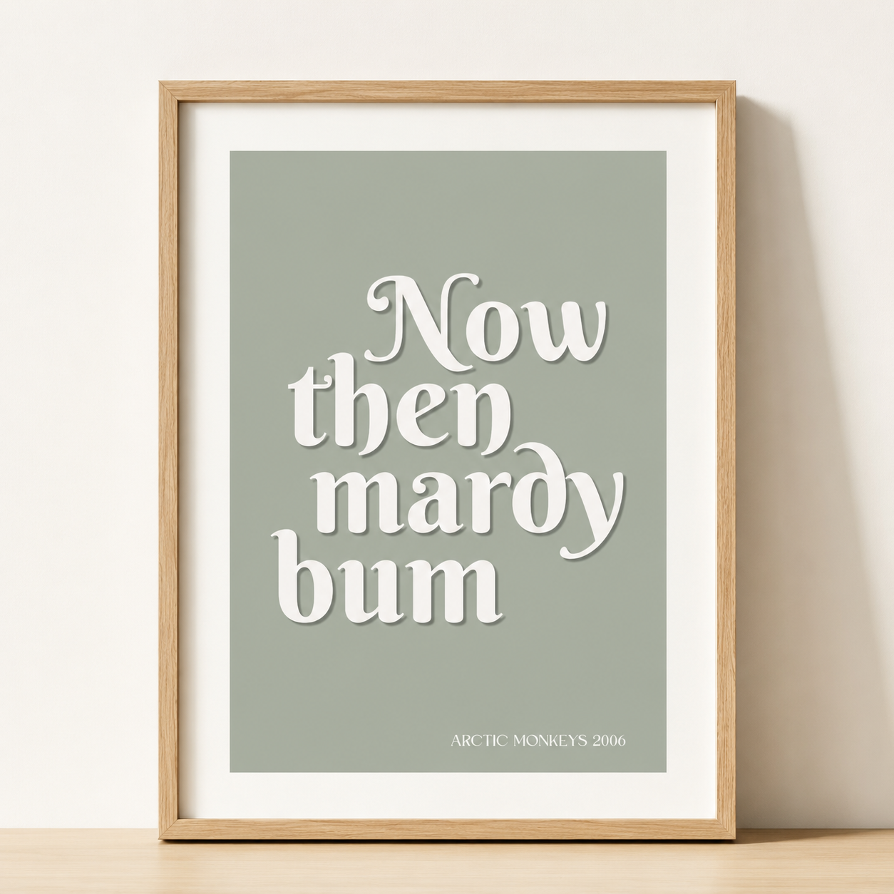 Sage green Arctic Monkeys "Now Then Mardy Bum" typography print in a natural oak frame with a white mount against a neutral wall.