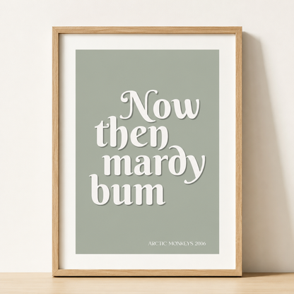 Sage green Arctic Monkeys "Now Then Mardy Bum" typography print in a natural oak frame with a white mount against a neutral wall.