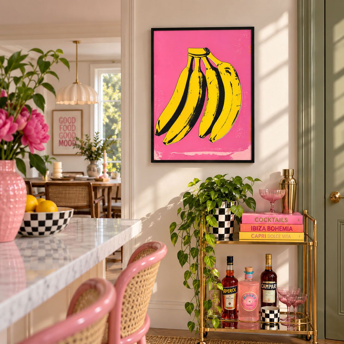 A framed wall art print featuring a bright pink background with bold yellow-and-black bananas, displayed in a styled kitchen or dining interior with plants, pink chairs, and a gold bar cart.