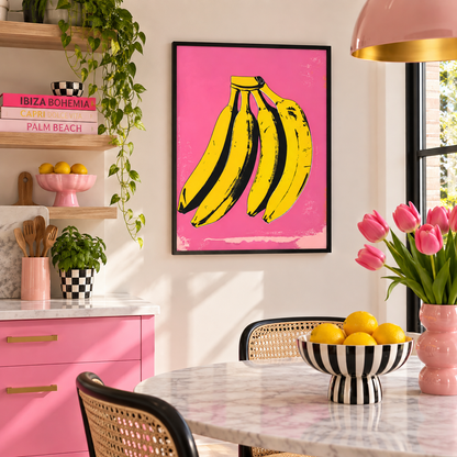 A framed pop-art style print featuring three yellow bananas on a bright pink background, displayed on a kitchen wall above a marble table with pink cabinets, flowers, and lemons.