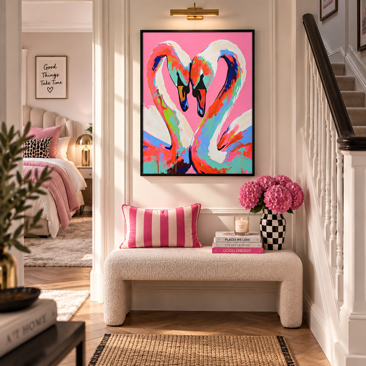 A colorful framed wall art print of two swans forming a heart shape, displayed in a bright, modern hallway above a white boucle bench with pink pillows and books.