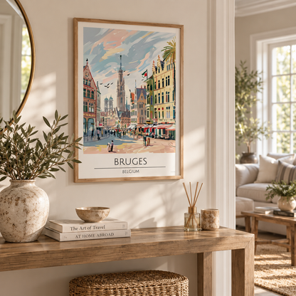 A framed travel poster of Bruges, Belgium, hanging on a light beige wall in a beautifully styled living room. The poster features a colorful watercolor-style illustration of the city's historic streets and architecture.