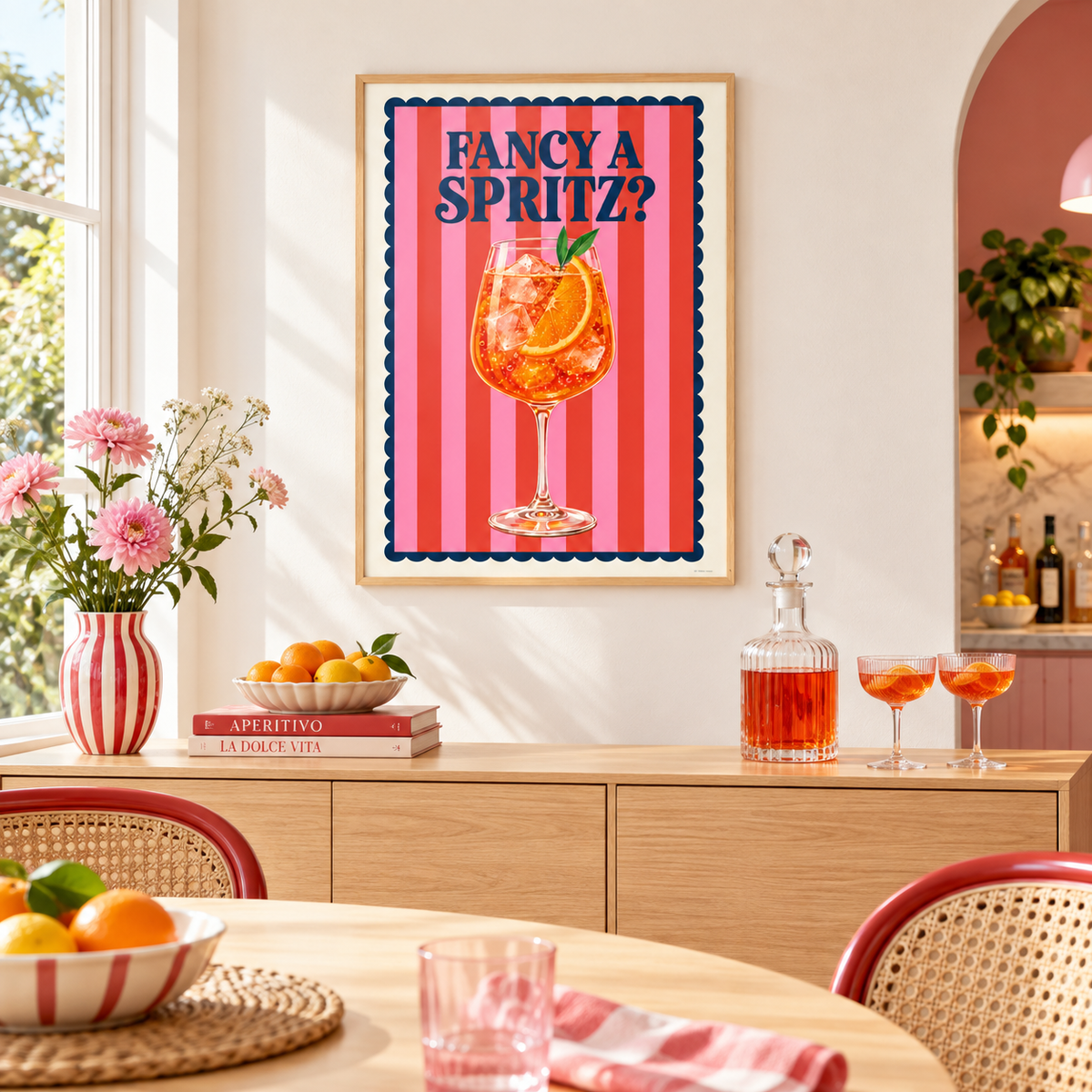A framed wall art print hanging above a wooden sideboard in a bright dining room. The print features the text 'FANCY A SPRITZ?' in navy lettering above an illustration of an orange cocktail in a glass, set against pink and red vertical stripes with a dark blue scalloped border.