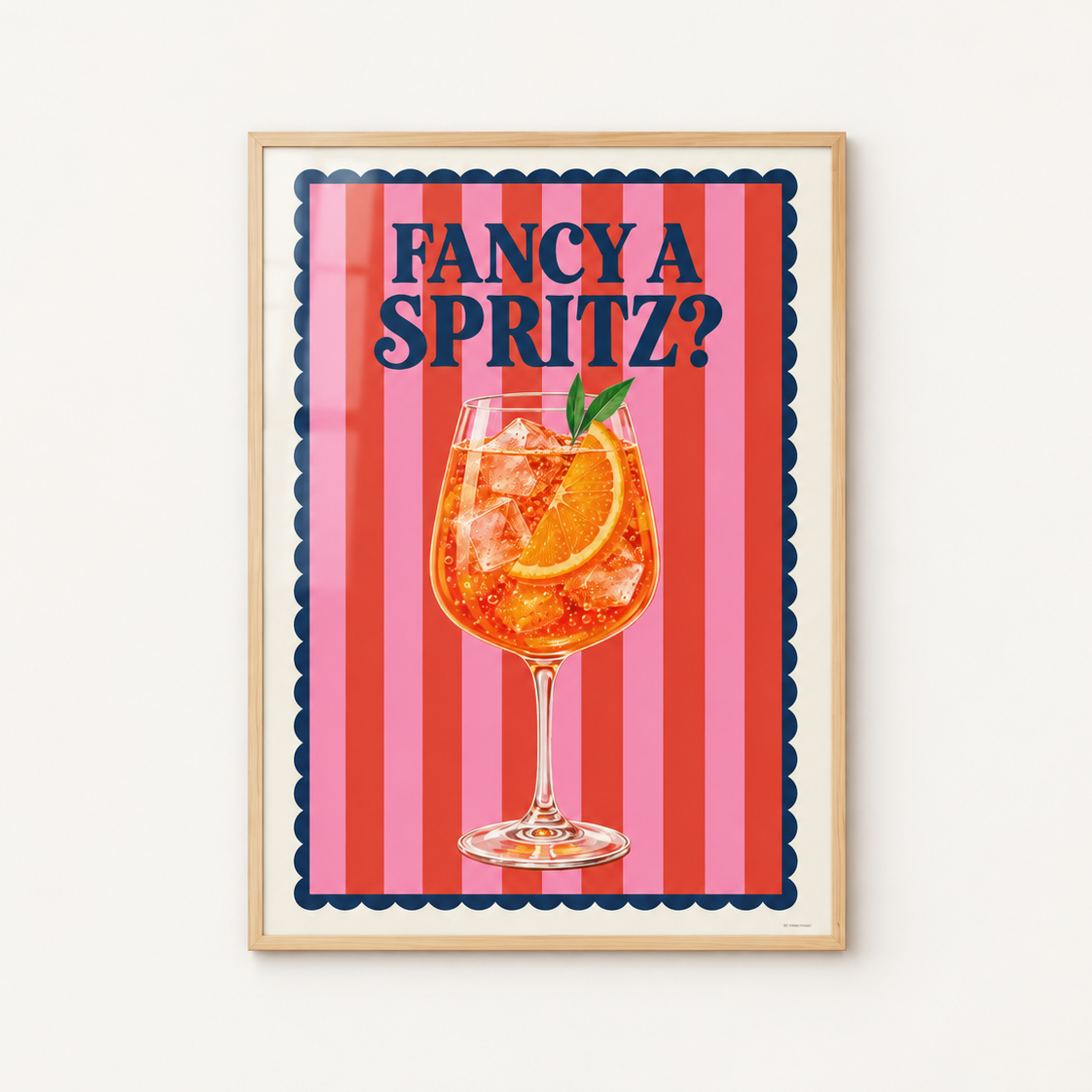 A framed art print featuring a cocktail illustration with the text 'FANCY A SPRITZ?' on a pink and red striped background, displayed on a light wall.