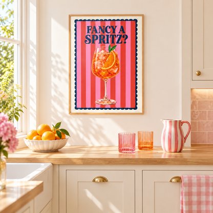 A framed wall art print with the text 'FANCY A SPRITZ?' hangs on a cream kitchen wall above a wooden counter. The print shows a pink striped background with a cocktail glass illustration and dark blue lettering.