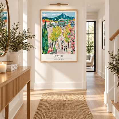 A colorful framed art print of Seoul, South Korea, hanging on a white hallway wall above a woven runner rug. The print features a vibrant, hand-painted style illustration of a scenic street with trees, mountains, and people, enclosed in a light wood frame.