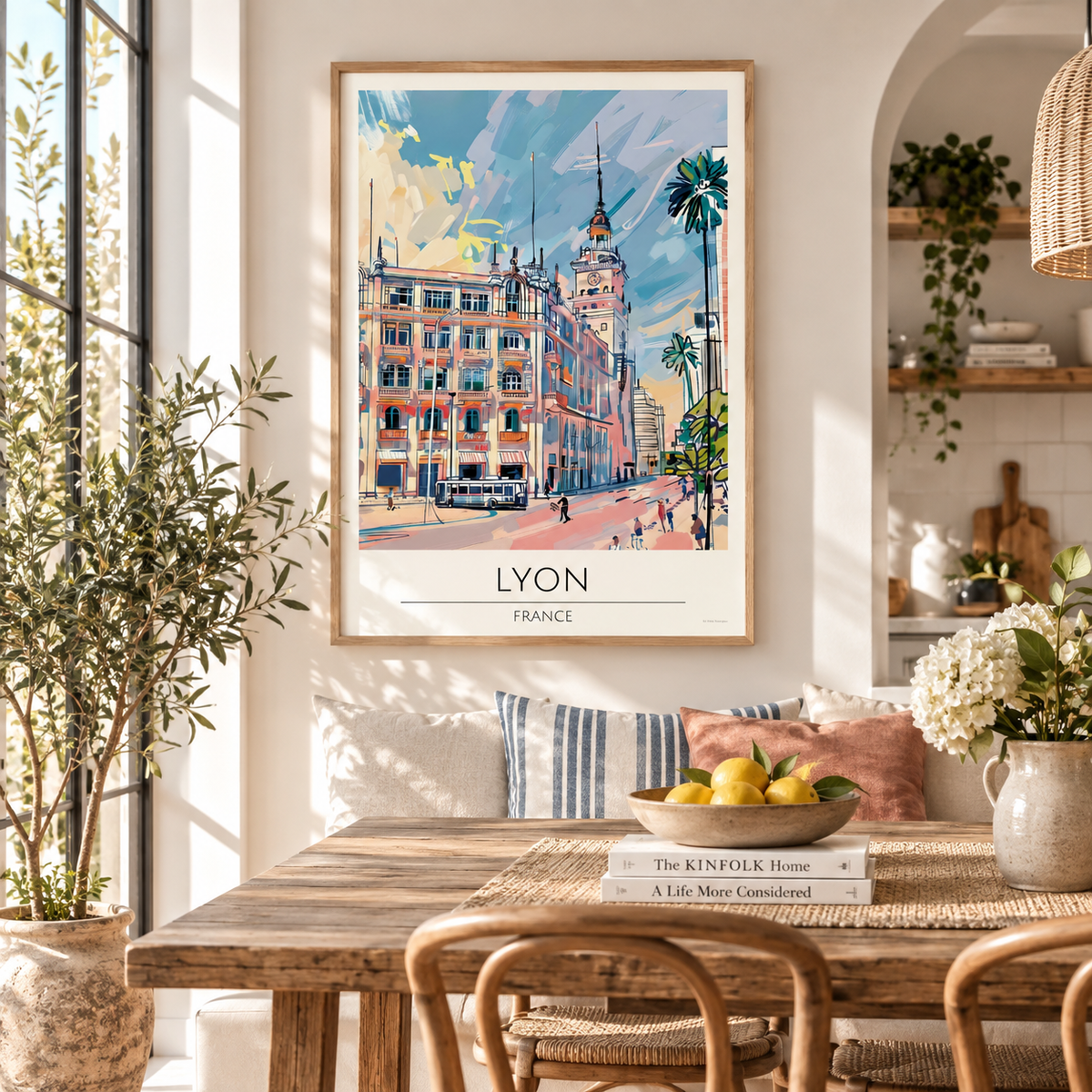 A framed art print of a colorful Lyon, France street scene hangs on a white wall in a bright, modern dining room with a wooden table, chairs, and decorative plants.