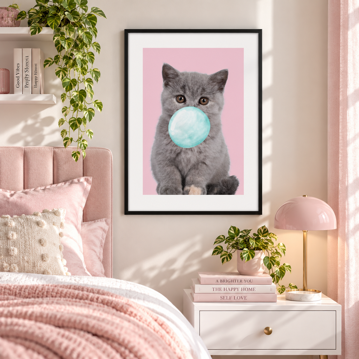 A framed wall art print featuring a gray kitten with a light blue gummy bubble on a pink background, displayed above a bed in a styled bedroom with pink bedding, a white nightstand, a pink lamp, and green plants.