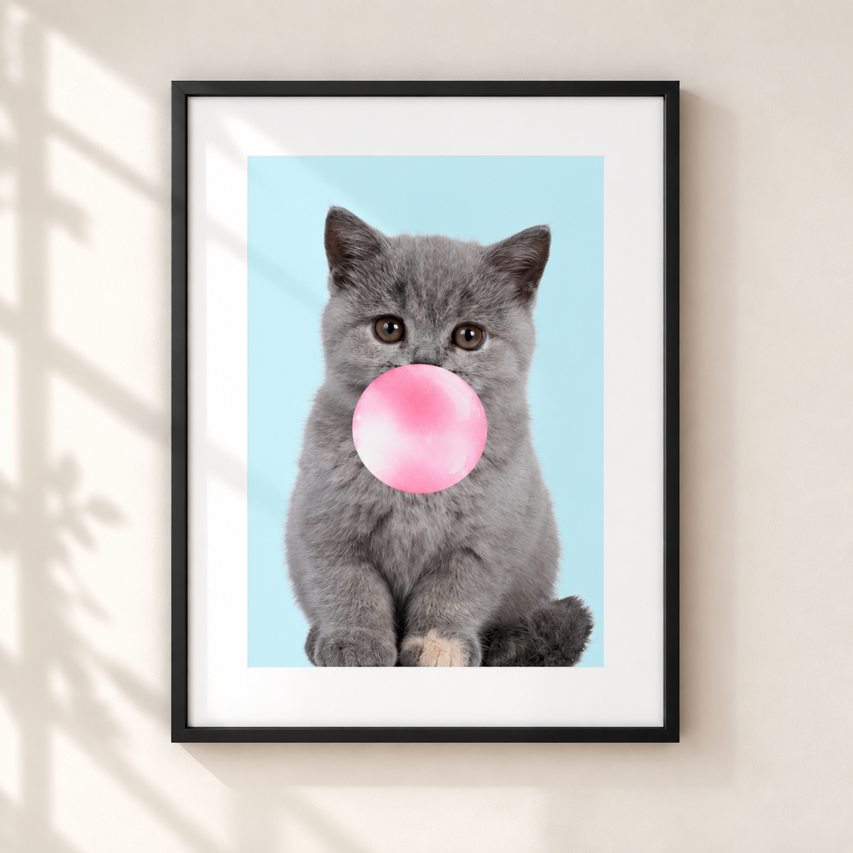 A framed art print featuring a gray kitten blowing a pink bubblegum bubble against a light blue background, displayed on a beige wall with soft shadow overlays.