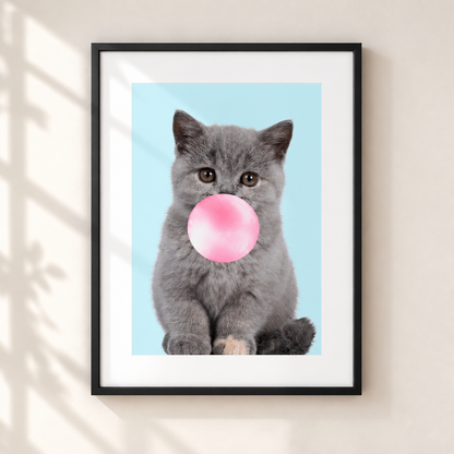 A framed art print featuring a gray kitten blowing a pink bubblegum bubble against a light blue background, displayed on a beige wall with soft shadow overlays.