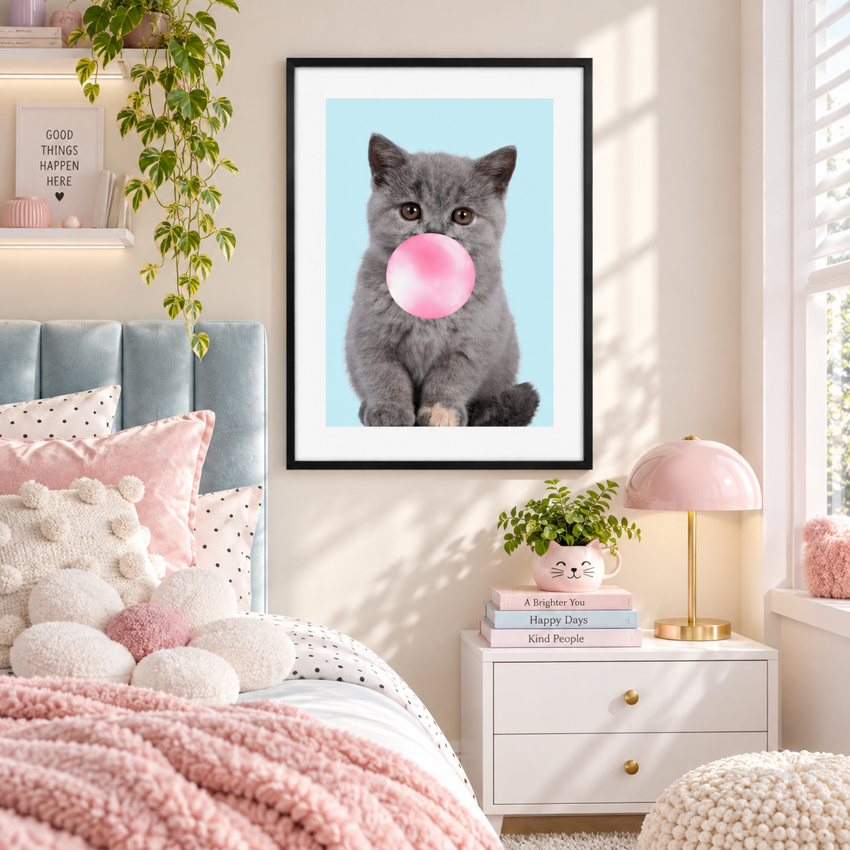 A framed wall art print showing a gray kitten blowing a pink bubblegum bubble against a light blue background, displayed above a bed in a styled bedroom with pink bedding, a white nightstand, and a pink lamp.