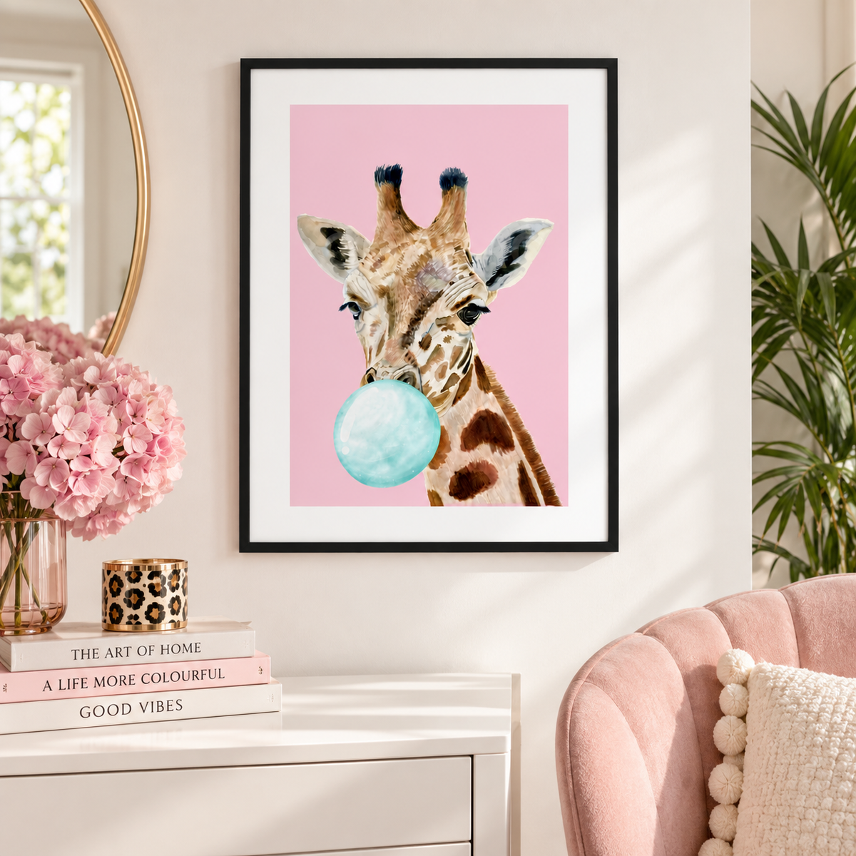 A framed art print featuring a watercolor-style giraffe blowing a light blue bubblegum bubble against a solid pink background, displayed on a white wall above a console table in a styled room.