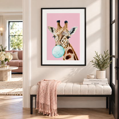 A framed art print featuring a giraffe blowing a light blue bubblegum bubble against a pink background, displayed on a wall above a tufted bench in a styled living room setting.