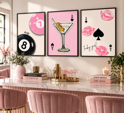 Three framed wall art prints with pink and black playing-card-inspired graphics hang above a marble counter in a styled interior. The prints feature a pink 1-ball, a martini glass, and pink lips with text including 'lady' and 'happy girl.