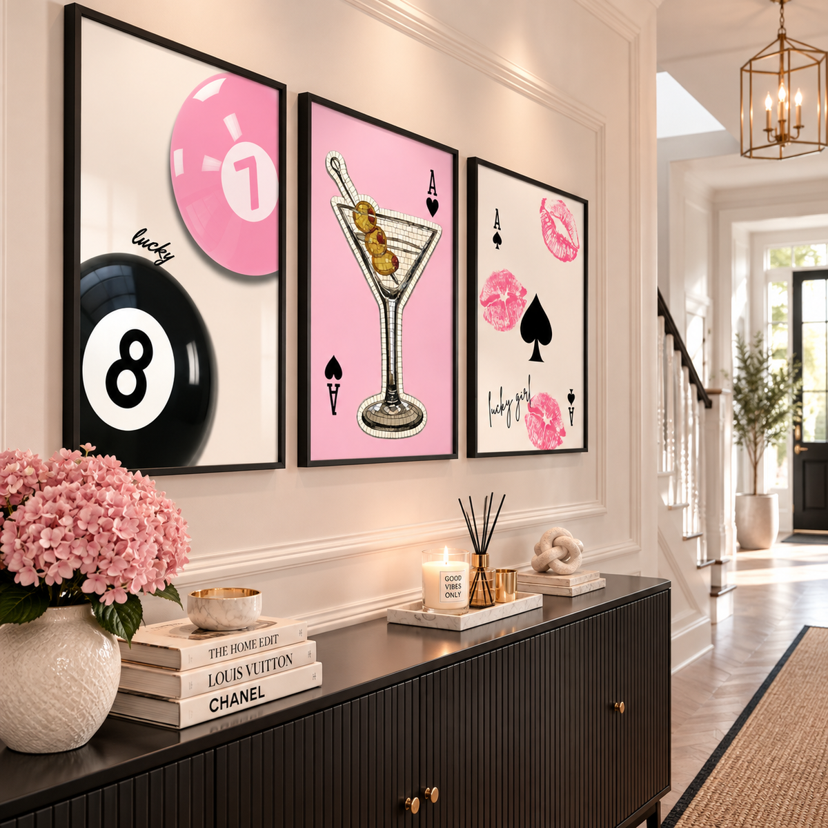 Three framed vertical wall art prints with pink and black gambling-themed illustrations are displayed on a white wall above a dark wood console table in a bright, modern hallway.