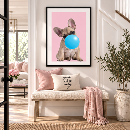 A framed wall art print featuring a light brown French Bulldog blowing a bright blue bubblegum bubble against a solid pink background, displayed in a modern, bright entryway above a cozy bench with pink pillows and a throw blanket.
