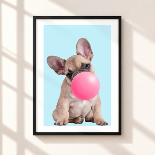 A framed art print featuring a cute French Bulldog puppy blowing a pink bubblegum bubble against a light blue background, displayed on a warm beige wall with soft window shadow overlays.