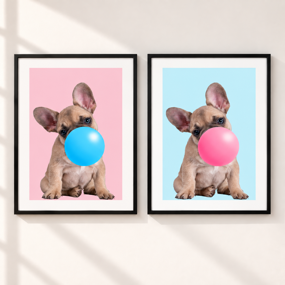 Two framed wall art prints hang side by side on a light beige wall. Each print shows a small tan dog with large ears blowing a large bubble: one bubble is bright blue on a pink background, and the other is pink on a light blue background.