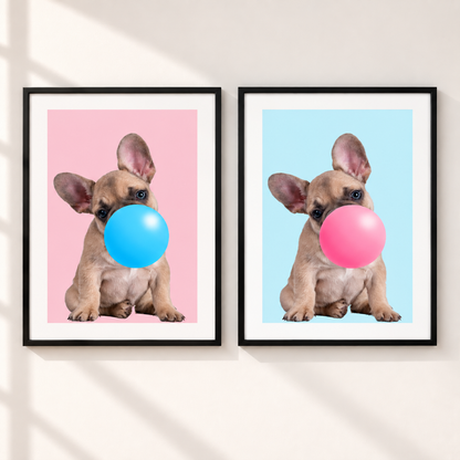 Two framed wall art prints hang side by side on a light beige wall. Each print shows a small tan dog with large ears blowing a large bubble: one bubble is bright blue on a pink background, and the other is pink on a light blue background.