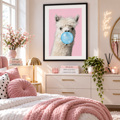 A framed wall art print featuring a fluffy white alpaca with a blue bubblegum bubble on a pink background, displayed in a styled bedroom setting above a white dresser.
