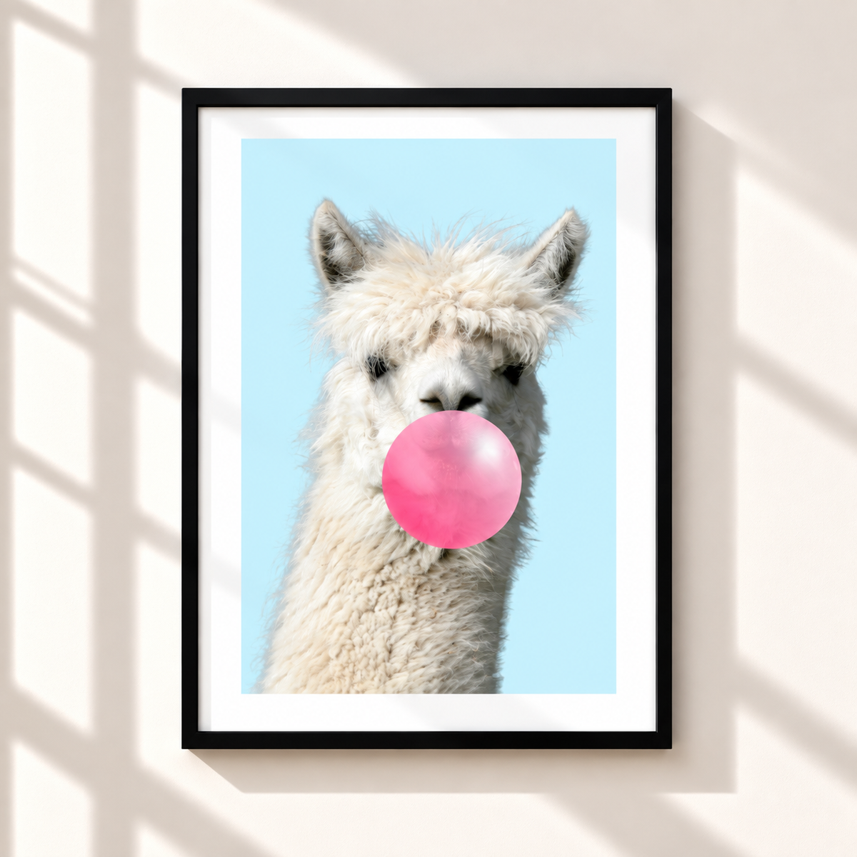 A framed art print featuring a close-up photograph of a fluffy white alpaca blowing a large pink bubblegum bubble against a light blue background, displayed on a beige wall with soft window shadow patterns.