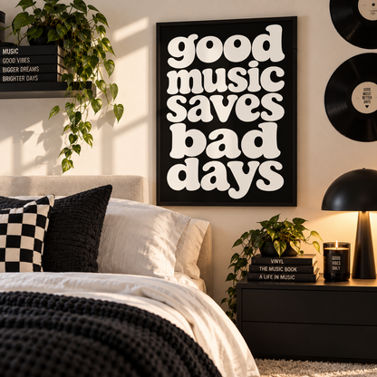 A framed wall art print with the phrase 'good music saves bad days' in large white letters on a black background, displayed above a bed in a styled bedroom setting with plants, pillows, a lamp, and vinyl records.