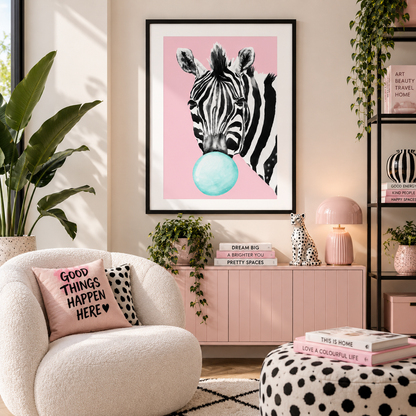 A framed wall art print featuring a black-and-white zebra blowing a light blue bubble, displayed in a styled living room above a pink cabinet. The room includes a cream boucle chair, potted plants, books, and a black shelving unit.