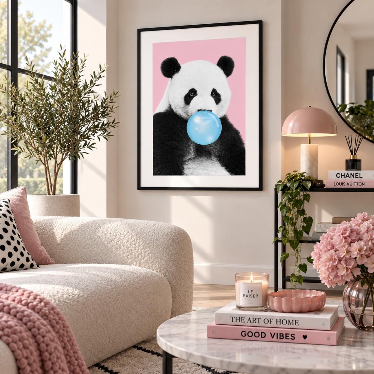 A framed art print featuring a black and white panda blowing a blue bubblegum bubble against a pink background, displayed on a beige wall in a modern, sunlit living room with a sofa, plants, and a marble coffee table.